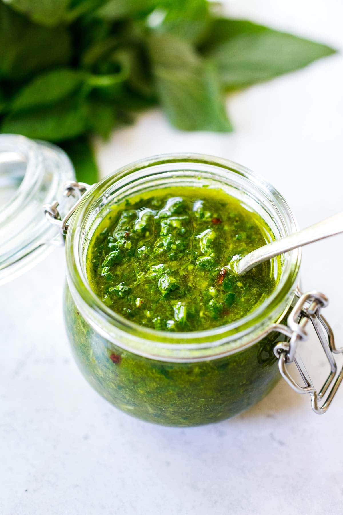 A jar of Basil Oil with a spoon. Made with fresh basil, garlic, olive oil, lemon, salt, and pepper, in just a few minutes.